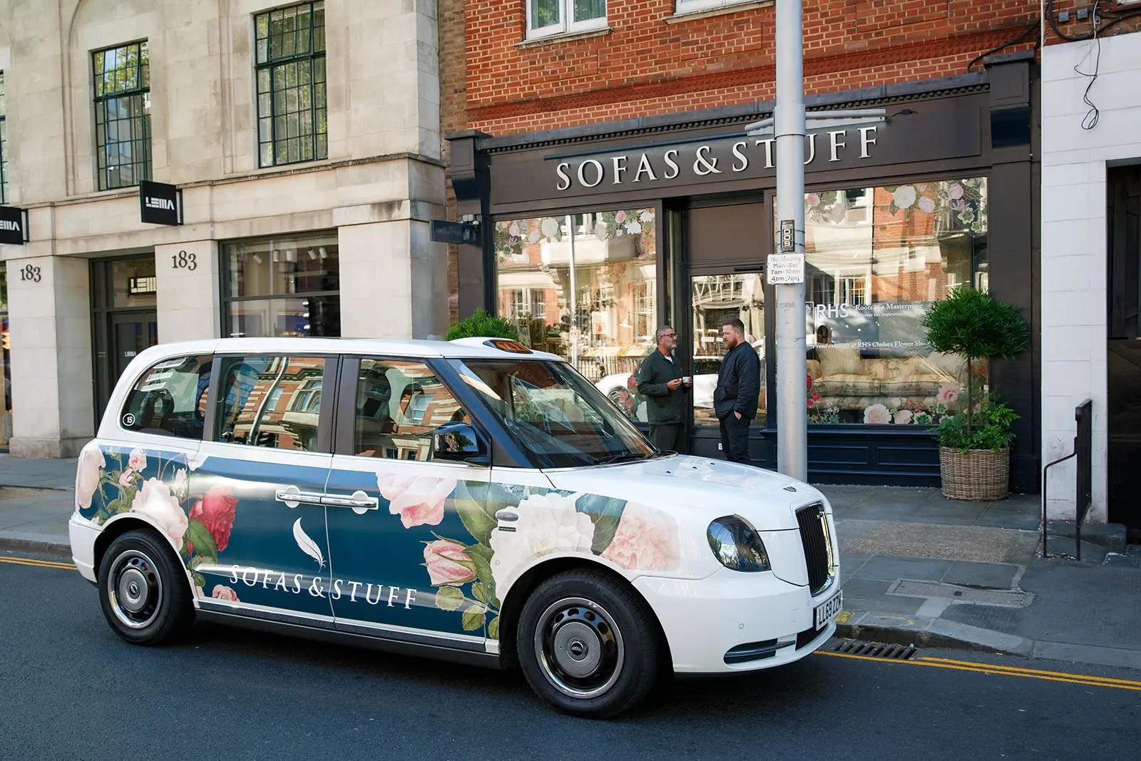 A branded Sofas & Stuff taxi with floral artwork parked outside a Sofas & Stuff store, extending the campaign visuals onto the street.